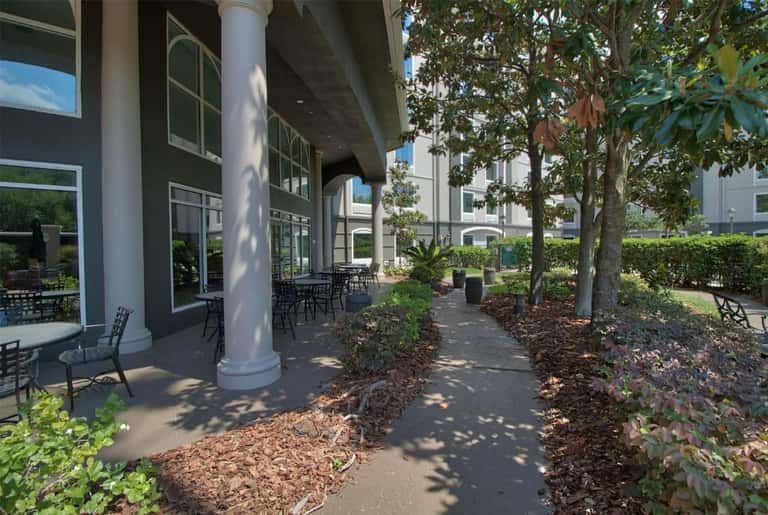 Covered outdoor walkway with columns, seating areas, and tropical landscaping alongside building