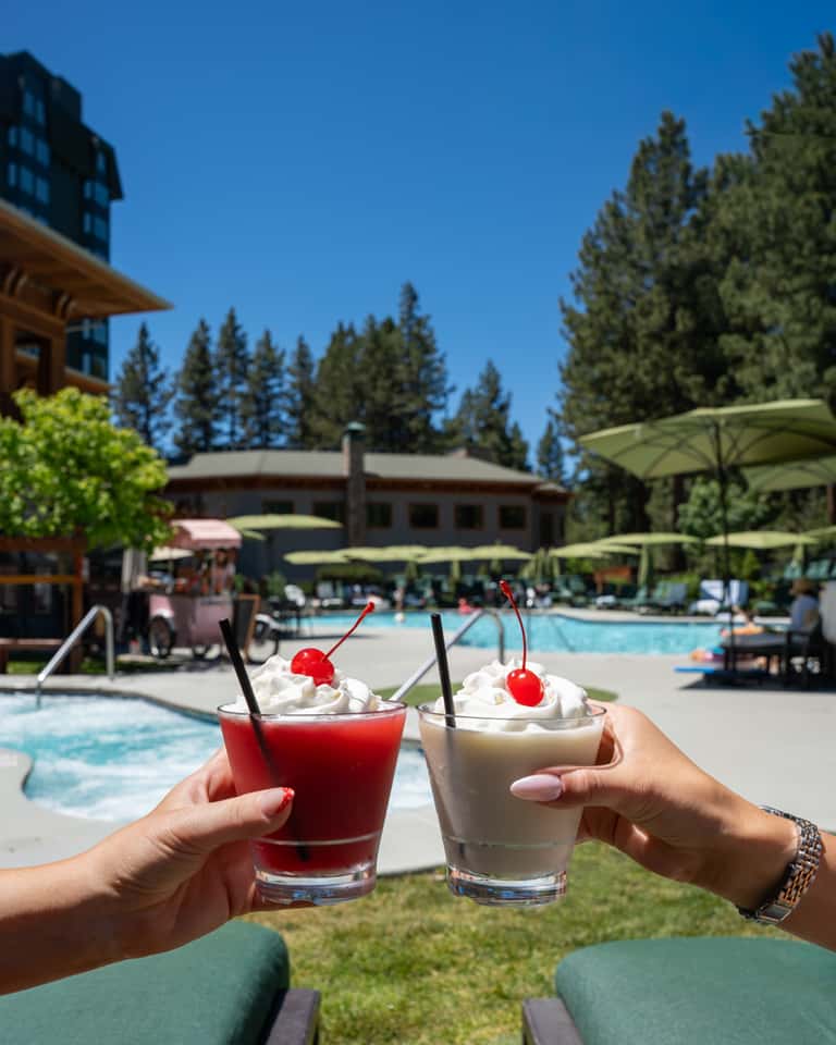 Two hands holding colorful cocktails with cherries near mountain resort pool, pine trees, and loungers