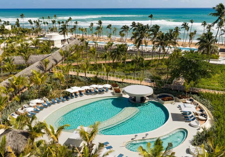 Aerial view of coastal resort pool with curved design, palm trees, beach access, and ocean backdrop