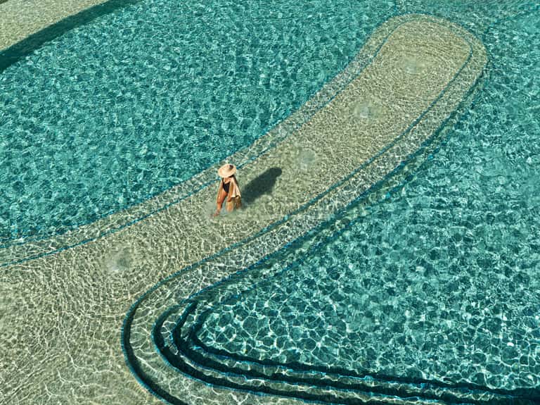 Aerial view of person wading in shallow turquoise water with sandy ripples visible from above