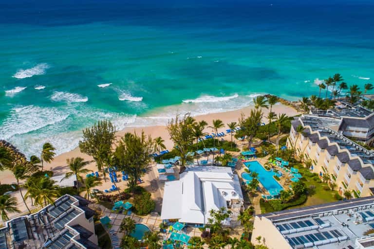 Aerial view of beachfront resort with pools, sandy beach, turquoise ocean waves, and multi-story hotel buildings