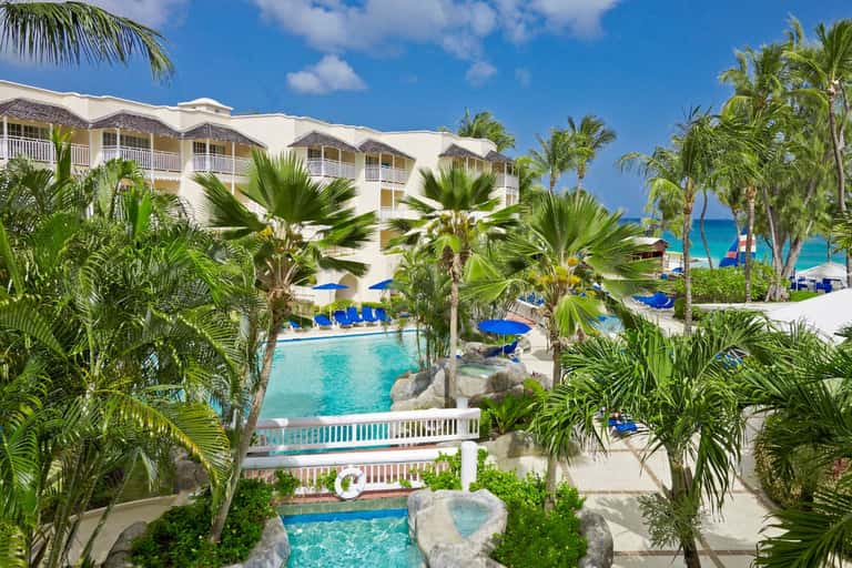 Beachfront resort with cream buildings, palm trees, pool, and blue umbrellas facing the ocean