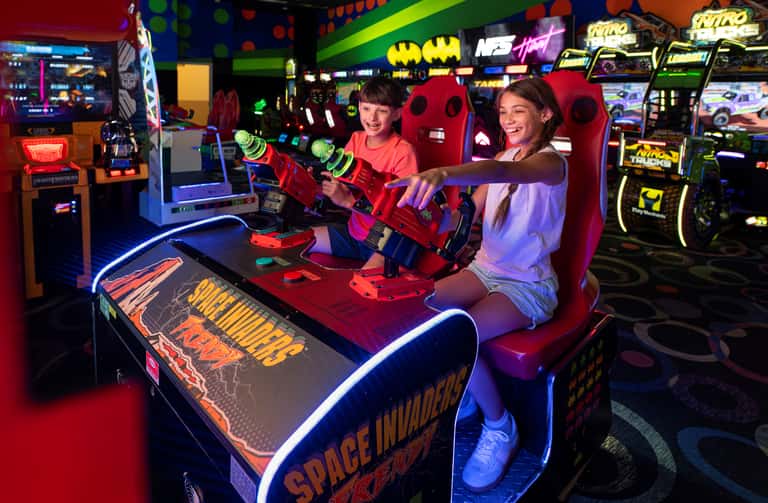 Two girls playing a red laser tag game in a colorful arcade filled with neon lighting and gaming machines