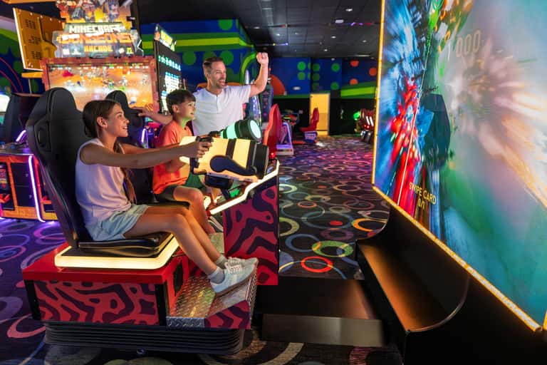 Family playing video racing game together in an arcade with neon decorations and colorful game cabinets