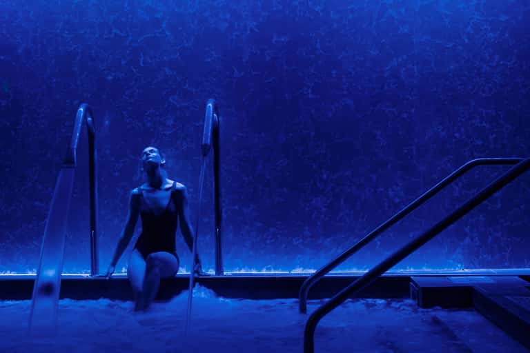 Woman in swimwear on pool steps at night with deep blue water and textured pool walls illuminated
