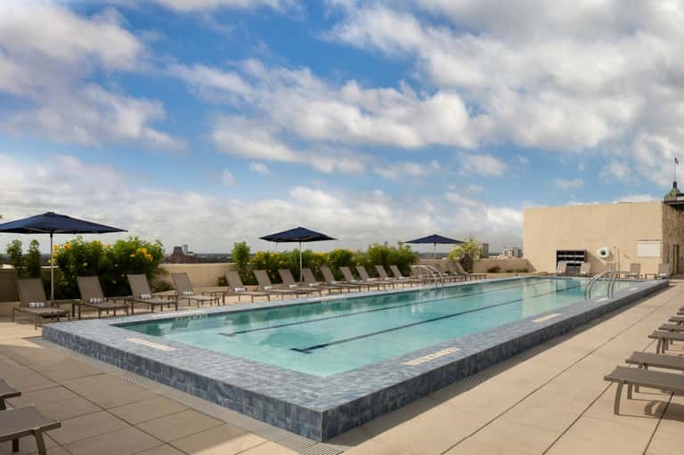 Rooftop lap pool with blue umbrellas and loungers overlooking a city skyline under cloudy skies