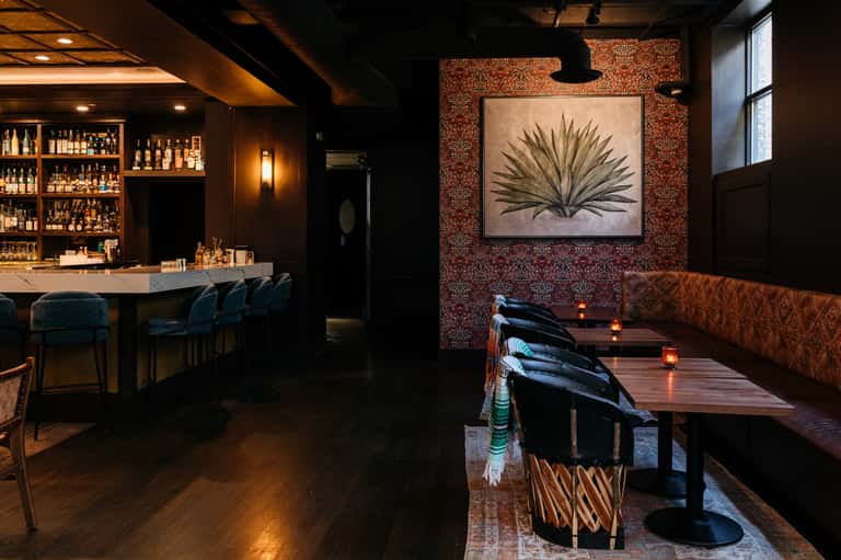 Intimate bar lounge with dark wood paneling, textured accent wall, artwork, dining tables, and ambient lighting