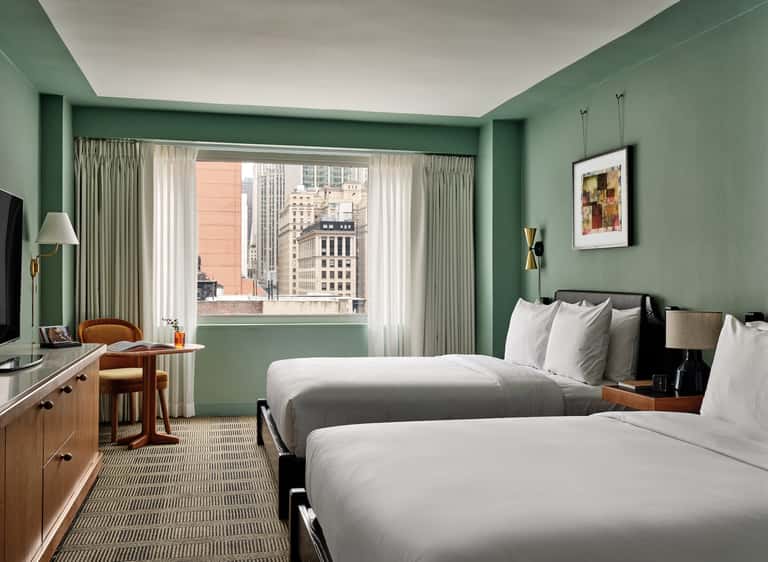 Hotel bedroom with sage green walls, two queen beds, wooden furniture, and cityscape window views