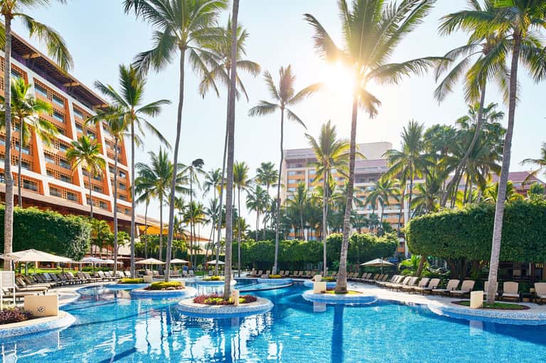 Luxurious resort pool with palm trees, lounge chairs, and orange-roofed buildings under bright sunlight