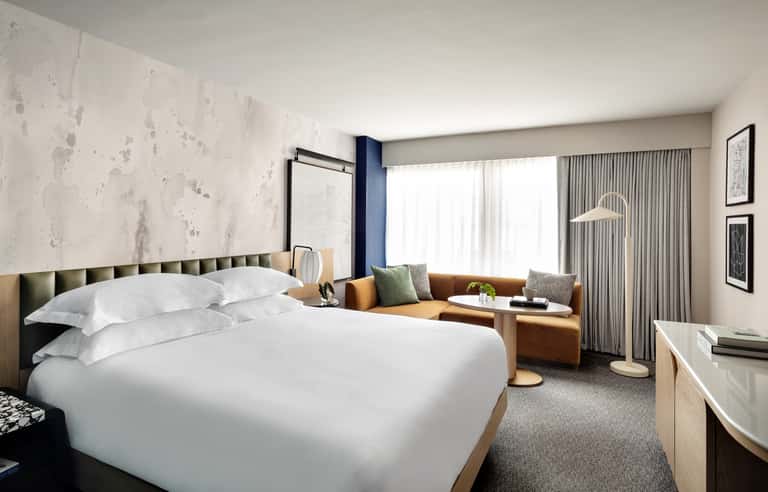 Contemporary hotel bedroom with marble accent wall, plush bedding, cognac sofa, and city views