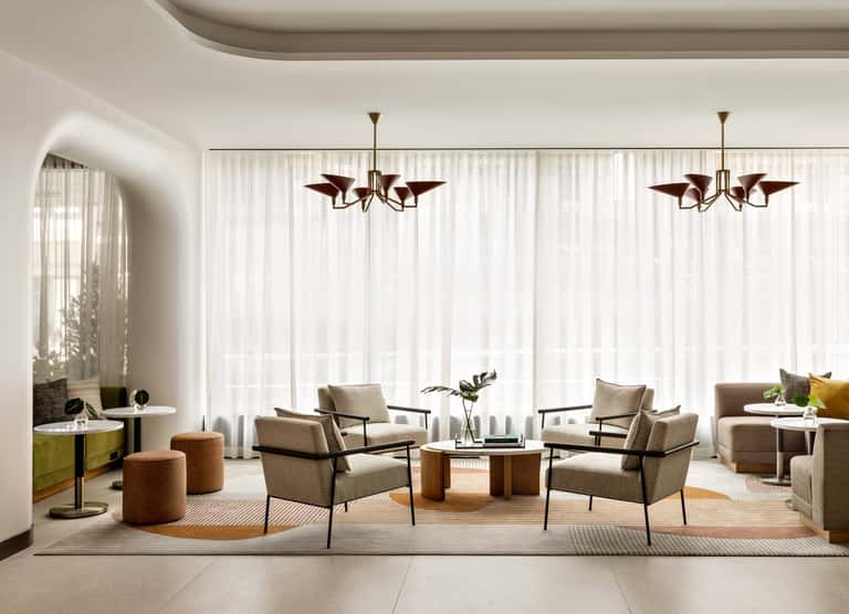 Modern luxury lobby lounge with neutral tones, beige chairs, wooden tables, and two brass pendant chandeliers above floor-to-ceiling windows.