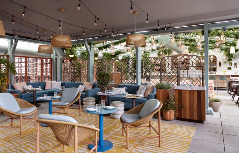 Outdoor rooftop lounge with blue seating, woven pendant lights, patterned rugs, and lattice privacy screens surrounded by greenery.