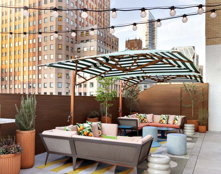 Urban rooftop terrace with striped pergola, string lights, coral sofas, wooden planters, and city skyline views above wooden privacy fence.