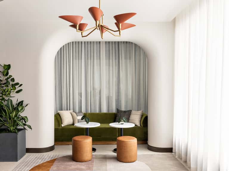 Curved nook seating area with olive green sofa, two tan ottoman poufs, white tables, abstract wall art, and coral mid-century chandelier.
