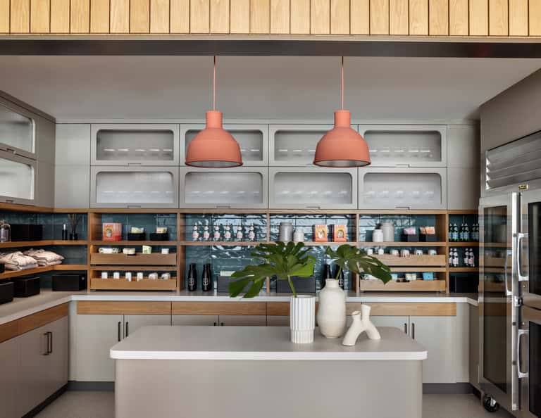 Modern café interior with coral pendant lights, open shelving with products, wooden accents, white countertop island, and professional kitchen setup.