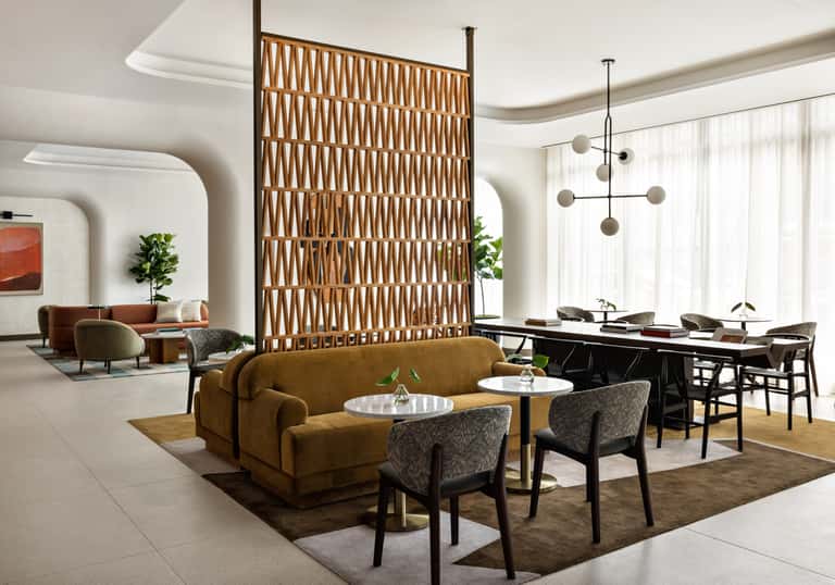 Contemporary hotel lounge with caramel sofa, patterned chairs, wooden room divider, black work desk, and sculptural pendant light fixture.