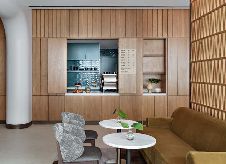 Modern beverage station with integrated wooden cabinetry, glass service window, white marble tables, tan seating, and geometric wood screens.