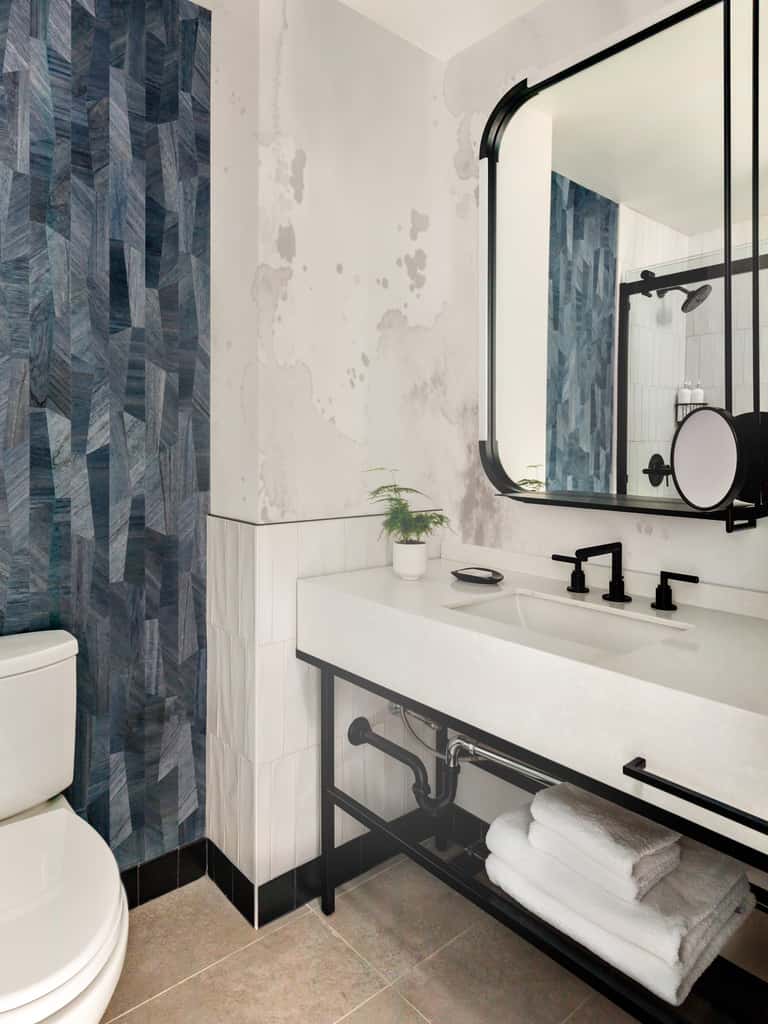 Contemporary bathroom with geometric blue tile shower wall, white vanity, black framed mirror, and black metal shelving unit.