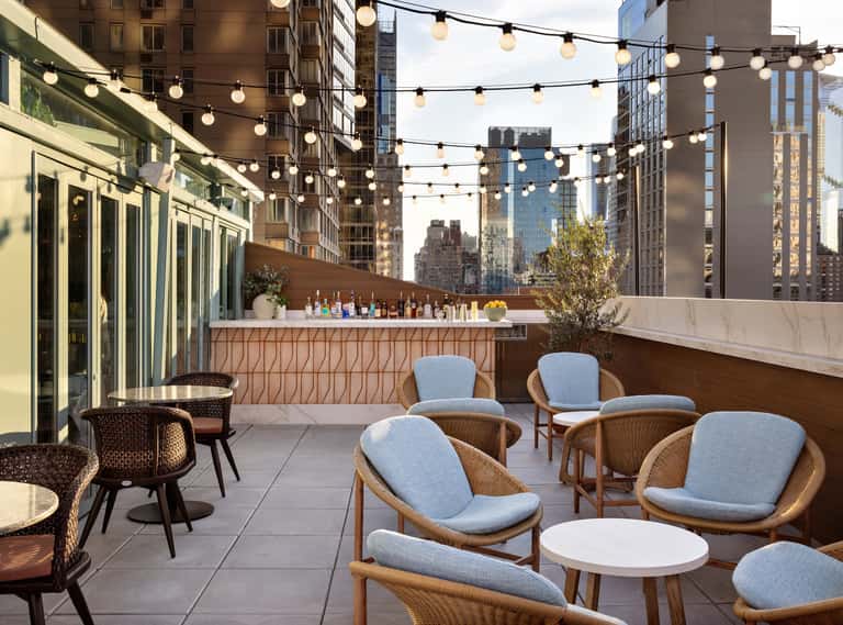 Rooftop terrace with string lights, wooden chairs with blue cushions, cityscape views, and bar counter with bottled beverages.