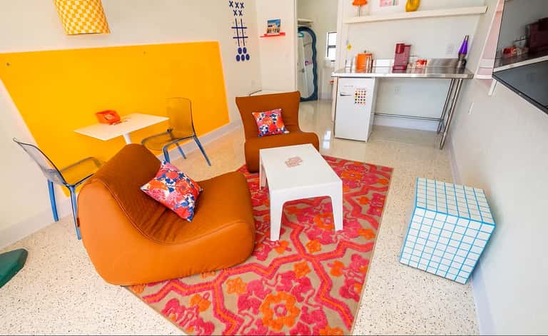 Colorful modern lounge with orange bean bag chairs, red patterned rug, yellow accent wall, and contemporary furnishings