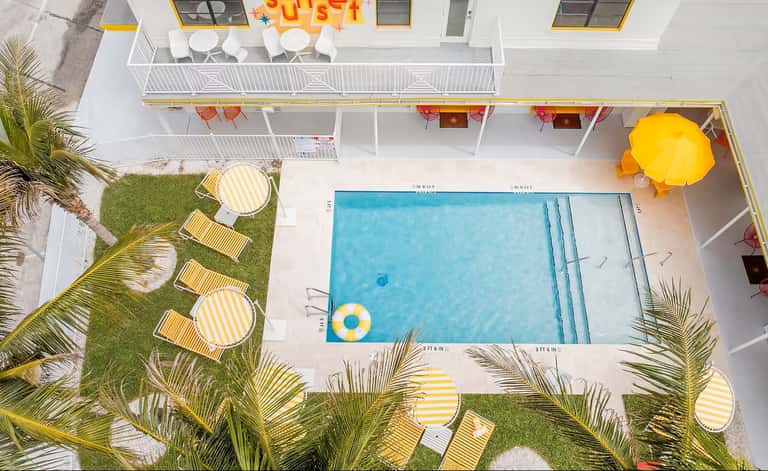 Aerial view of resort pool with striped lounge chairs, palm trees, and yellow umbrellas on sunny day