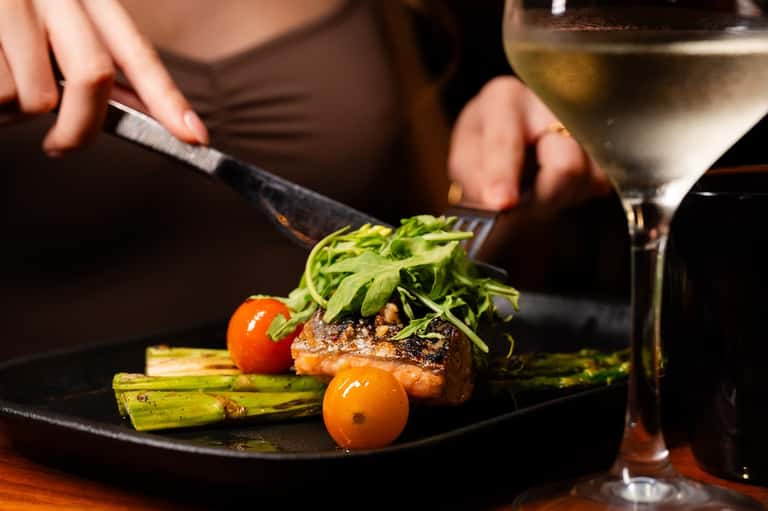 Close-up of a gourmet plated dish featuring seared meat, green asparagus, cherry tomatoes, and microgreens with wine glass