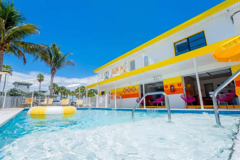 Vibrant resort pool with yellow and white floaties, pink metal chairs on deck, and colorful striped umbrellas
