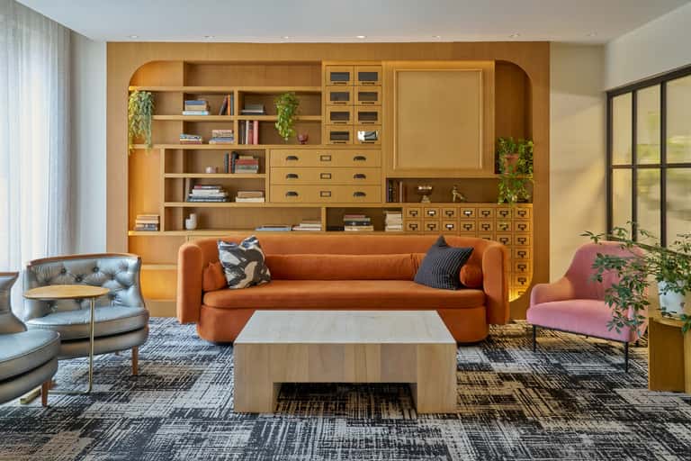 Contemporary hotel lounge with orange sofa, wooden shelving unit, blue accent chair, light wood flooring, and hanging plants