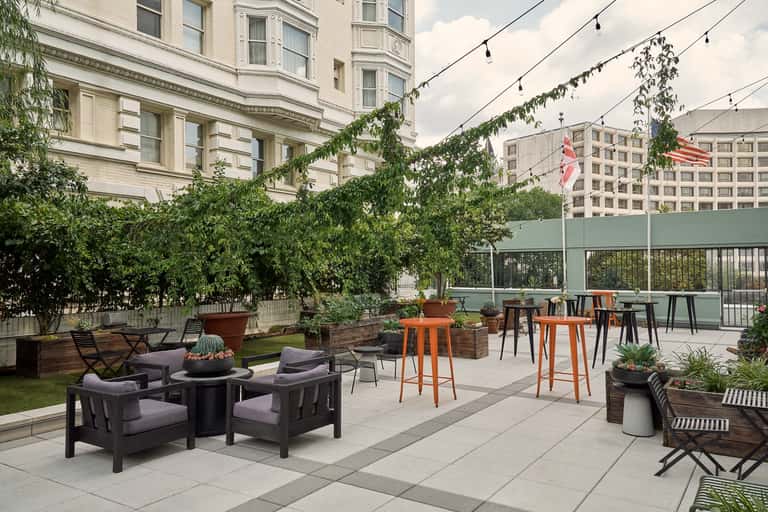 Upscale rooftop lounge with purple chairs, ivy-covered walls, orange side tables, string lights, and elevated city views