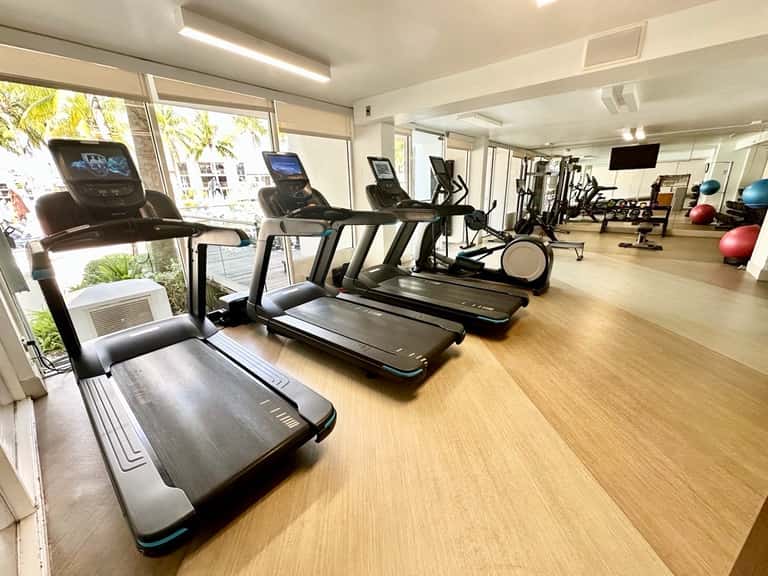 Modern fitness center with multiple treadmills, elliptical machines, and exercise equipment along bright windows overlooking palm trees