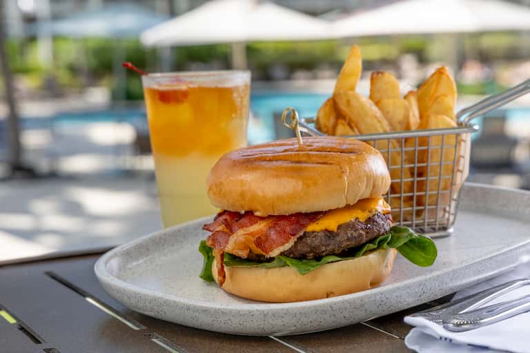 Gourmet bacon cheeseburger with fries and cocktail served poolside at resort restaurant with umbrellas visible in background