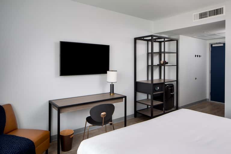 Modern hotel room with white walls, dark desk with TV, black industrial shelving unit, tan accent chair, and minimalist decor