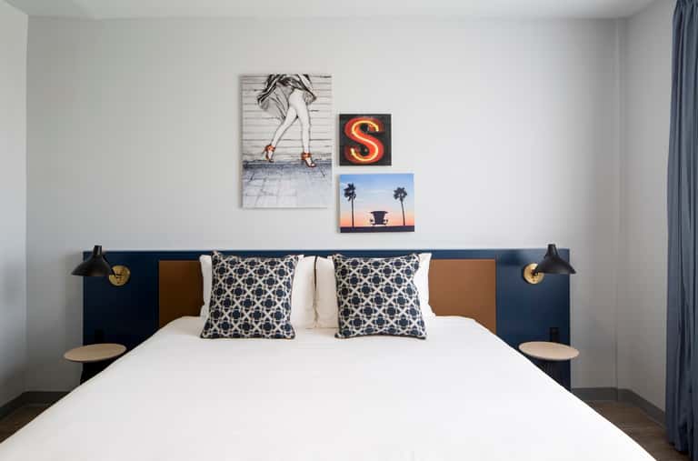 Contemporary bedroom with white bedding, patterned pillows, navy headboard with tan side panels, and pop art above bed