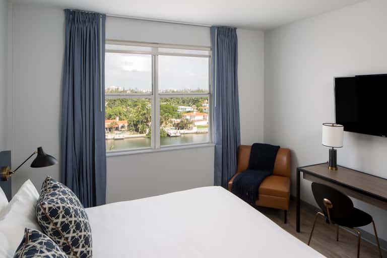 Bright hotel room with large windows overlooking water, blue curtains, white bed with patterned pillows, and tan accent chair