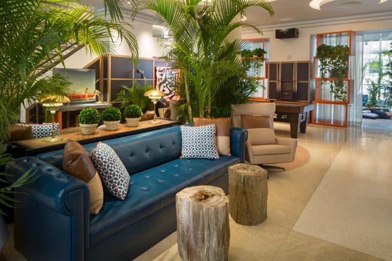 Upscale hotel lobby with blue velvet sofa, tropical plants, wooden accents, patterned pillows, and warm ambient lighting