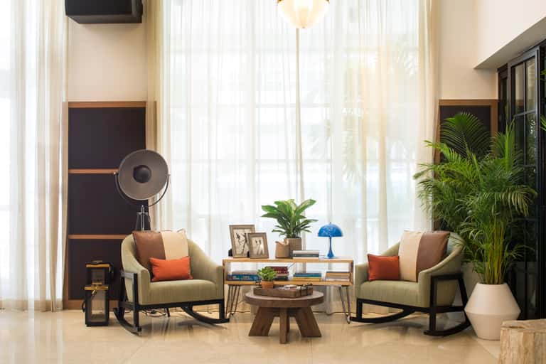 Luxury lounge with two sage green rocking chairs, wooden coffee table, potted plants, and floor-to-ceiling windows with sheer curtains