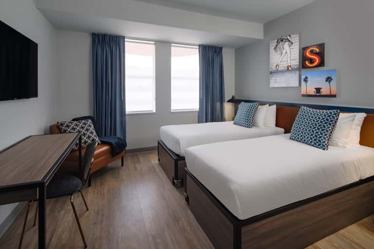 Modern hotel bedroom with two queen beds, blue curtains, wooden desk, and wall-mounted TV with contemporary artwork