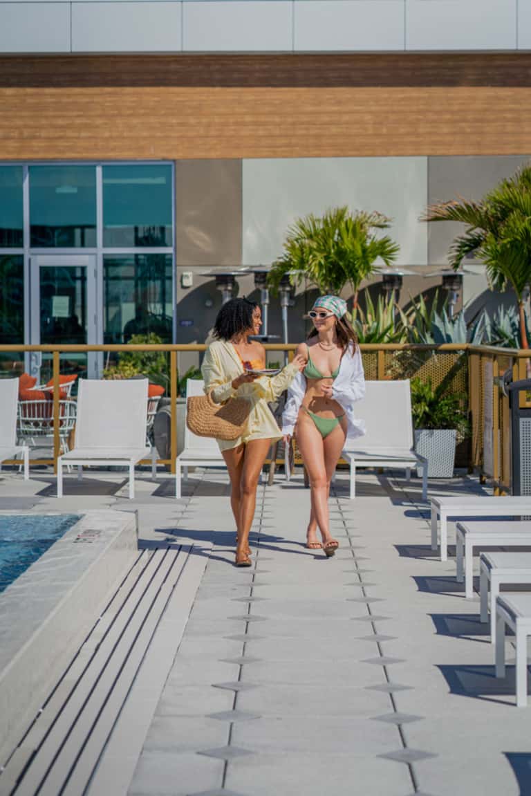 Two women walking on a modern resort deck with lounge chairs, pool, and contemporary building with wood accents