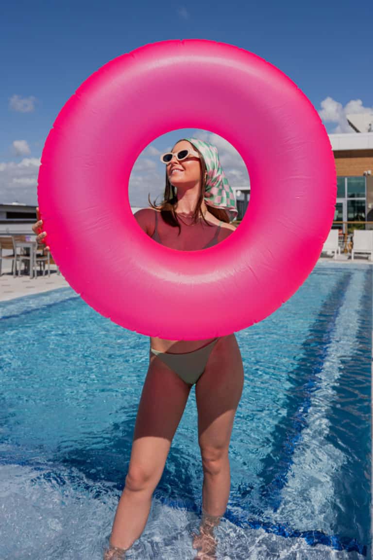 Woman in pool holding large pink inflatable ring float, wearing sunglasses and headscarf