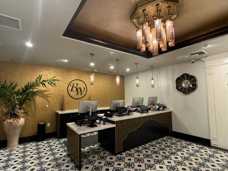 Elegant hotel reception desk with gold accent wall, geometric floor tiles, pendant lighting, and potted plants