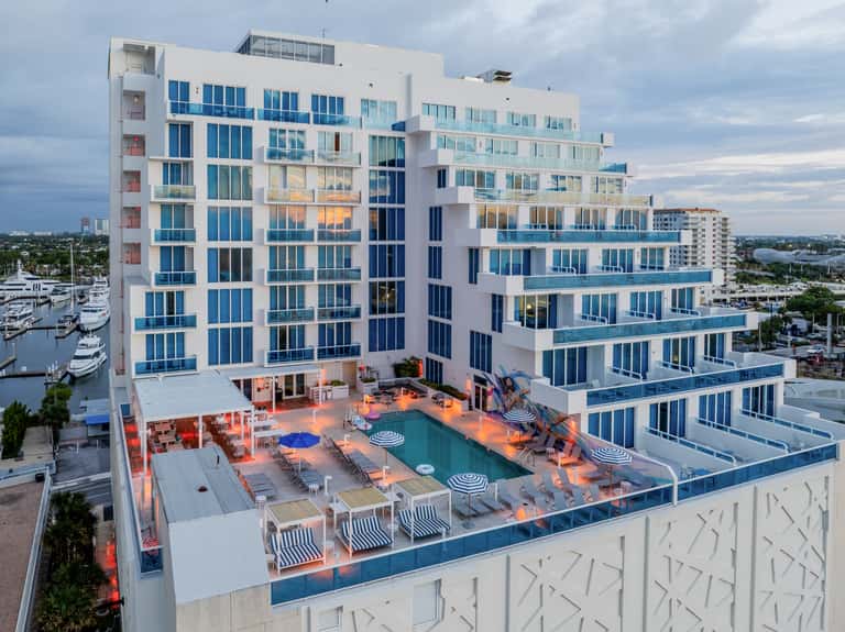 Modern waterfront hotel with blue and white facade, rooftop pool, lounge area, and marina views at dusk