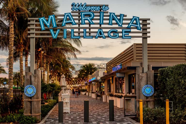 Marina Village neon sign entrance with palm trees, shops, and pedestrian walkway at dusk