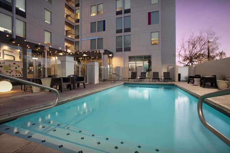Resort outdoor pool at dusk with illuminated modern building, lounge chairs, and purple twilight sky