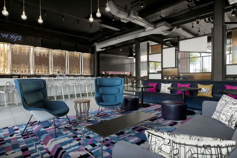 Modern lobby lounge featuring blue accent chairs, geometric patterned carpet, and elegant bar area