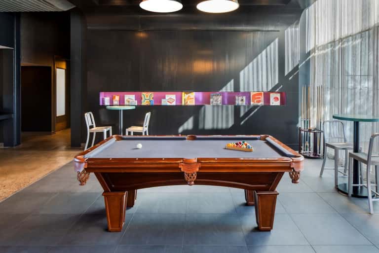 Modern recreation room with wooden billiards table, colorful digital art display, and floor-to-ceiling windows with natural light