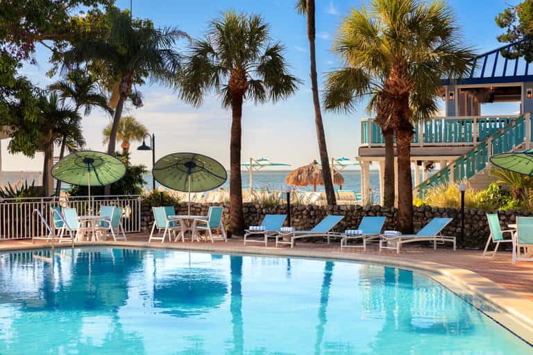 Resort pool area with blue lounge chairs, green umbrellas, palm trees, and ocean view