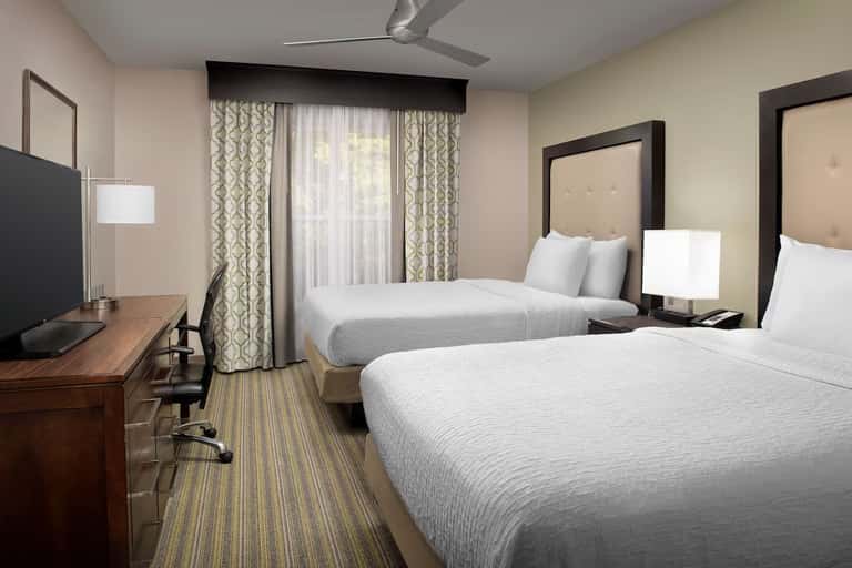 Hotel guest room with two queen beds, work desk, flat-screen TV, and patterned curtains overlooking balcony
