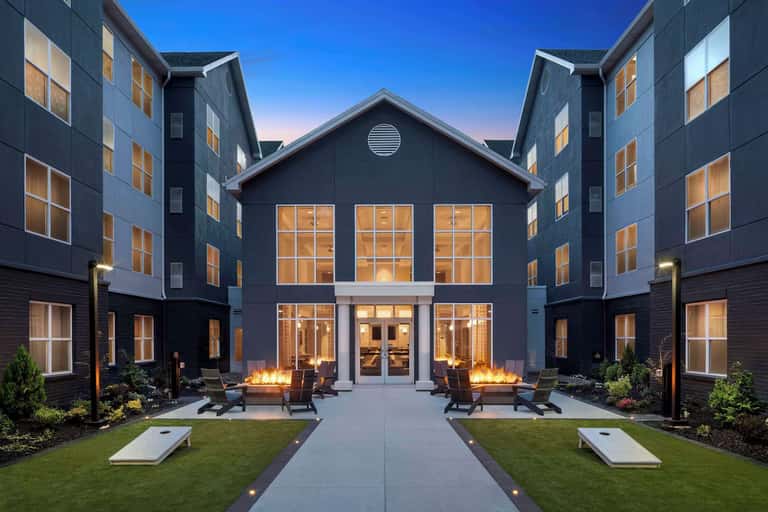 Modern multi-story hotel exterior at dusk with illuminated windows, fire pits, and manicured courtyard