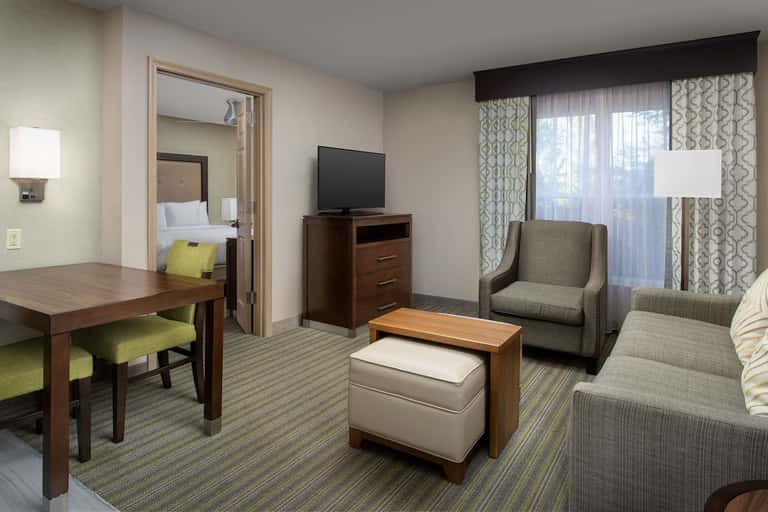 Hotel suite living area with wood furnishings, gray seating, TV, desk, and bedroom visible through doorway