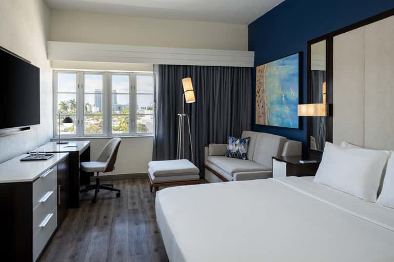 Modern hotel room with king bed, work desk, sitting area, dark curtains, and city skyline views through large windows